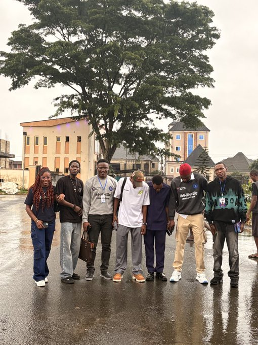 A group of eight people stand together posing for a photo in front of a large tree and building on a wet, overcast day.