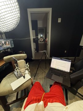 A first-person view of someone in a Santa Claus costume sitting at a desk with a laptop and microphone in what appears to be a recording studio setup with sound equipment visible.