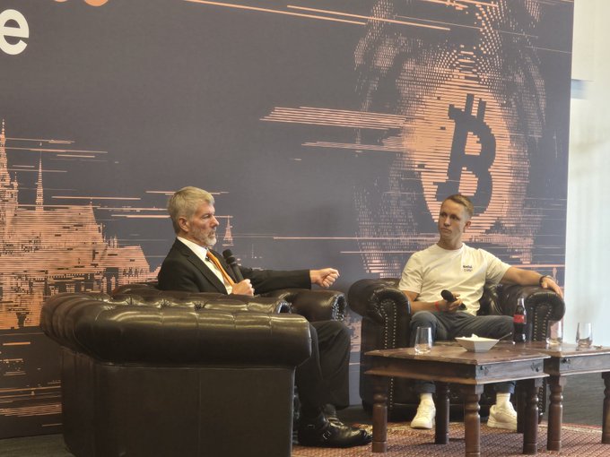 The image features a man sitting in a chair, wearing a suit and tie, while another person sits across from him on a couch. They are both facing each other, engaged in conversation. The background of the scene includes a large mural depicting a Bitcoin symbol, suggesting that they might be discussing cryptocurrency or related topics. There is also a bottle placed between them, possibly containing a beverage to enjoy during their discussion.