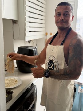 A casual photo of a smiling man in a kitchen wearing an apron, giving a thumbs-up while holding up a piece of cooked food, with a plate of food on the counter beside him.
