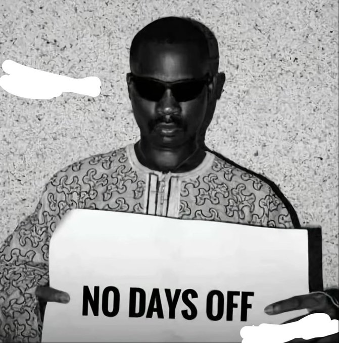 A black and white image of a man wearing sunglasses and a patterned traditional shirt, holding a sign that reads "NO DAYS OFF".