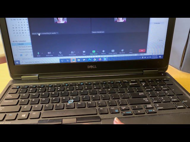 The image shows an open laptop computer with a person's hand pressing a key on the keyboard. The laptop screen displays two faces, possibly from a video call or a presentation. The background of the image also includes a calendar and a book, suggesting that this might be taking place in a workspace or home office setting.