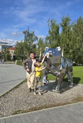 A woman posing next to a Transformers Bumblebee statue outdoors on a sunny day with green trees in the background.