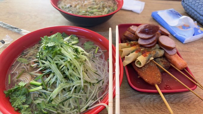 A meal of two bowls of noodle soup with cilantro and a side plate of skewers and sliced sausage, accompanied by a pack of tissues.