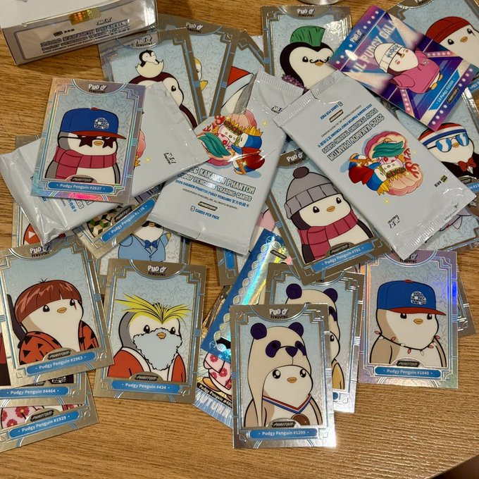 A collection of Pudgy Penguins trading cards in sealed foil wrappers scattered on a wooden table, featuring various penguin characters like Pudgy Penguin #2837, #4044, #3627, #4238, and #4206 in winter clothing and hats.