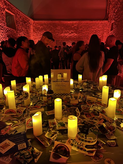 A dimly lit memorial event features a table covered in candles, stickers, and a sign reading 'THE DEEPEST SACRIFICABLE,' with people standing in the red-lit background.