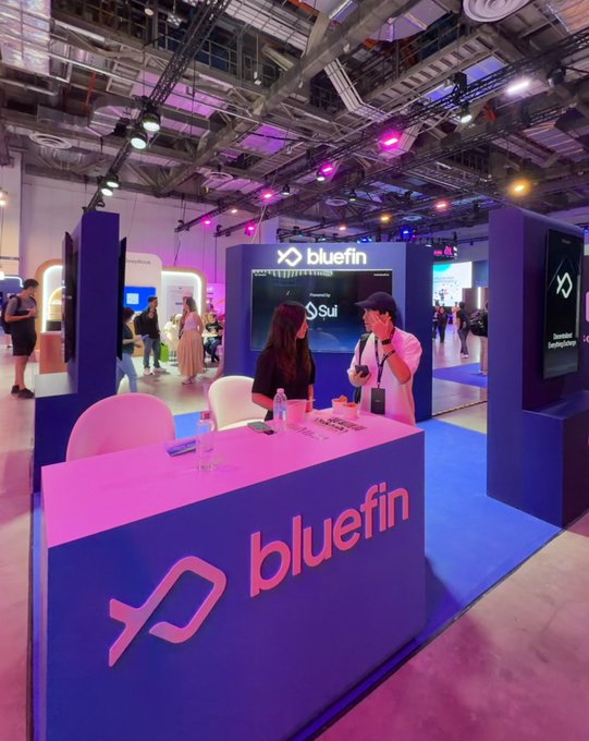 A vibrant Bluefin booth at Sui Basecamp's Day 1, featuring branded signage and a pink-lit counter where staff engage with attendees amid the bustling conference atmosphere, with screens displaying "bluefin" and "Sui" logos throughout the exhibit space.