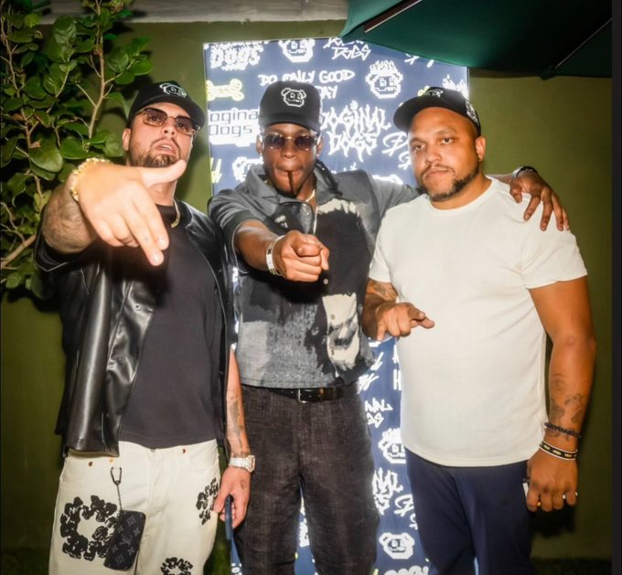 Three men pose together in front of a branded backdrop featuring "Doginal Dogs," "Do Only Good," and "Original Dogs" logos, with the central figure in a graphic skull shirt and sunglasses pointing at the camera. The image ties to the post by visually promoting the doginaldogs.com ecosystem and its expanding suite of NFT projects ahead of the next cycle.