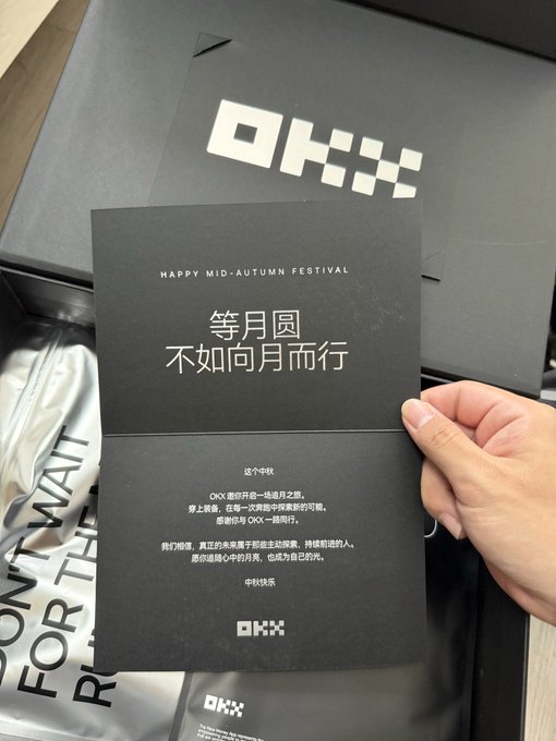 A hand holds a black OKX-branded Mid-Autumn Festival greeting card with Chinese text '等月圆 不如向月而行' above a branded gift box.