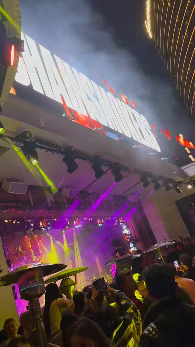 The image shows a large crowd of people gathered outside a venue, possibly attending an event or concert. The crowd appears to be enjoying themselves as they take photos with their phones. In the background, there are neon lights illuminating the area, creating a vibrant atmosphere. A prominent sign in front of the crowd reads "The Chainsmokers", indicating that this is likely a performance by the popular band "The Chainsmokers". The post also mentions that the event takes place at ApeCoin Vegas and is scheduled for early @DeblockApp investors.

There are several people standing around, some closer to the camera while others are further away in the background. They seem to be engaged in conversations or taking photos with their phones. Overall, it appears to be a lively scene filled with excitement and anticipation as fans gather outside the venue for this event.