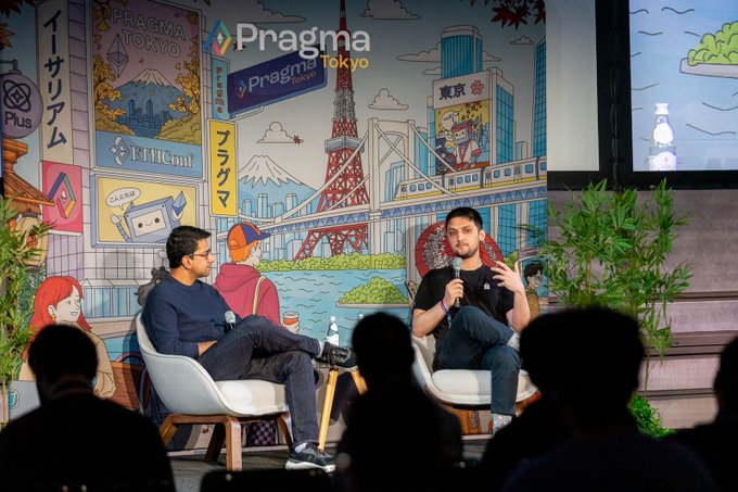 Two men sit on chairs on stage discussing at the 'Pragma Tokyo' event, with a colorful Tokyo-themed mural featuring Mount Fuji and Tokyo Tower behind them.