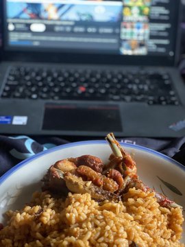A close-up of a plate of yellow rice with meat on a bone, positioned in front of an open laptop displaying video editing software.