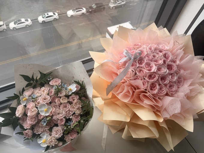 Two large bouquets of pink flowers, including roses wrapped in peach-colored paper, sit on a windowsill overlooking a street with passing cars.