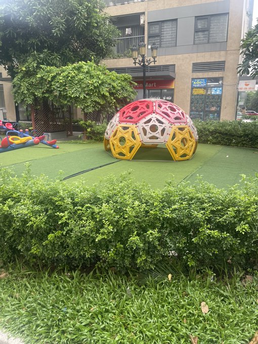 An outdoor playground scene featuring a colorful geodesic dome climber in red, yellow, and white on artificial green turf, with a building, trees, and shrubbery in the background.