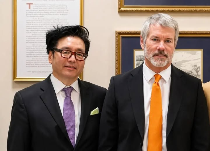 Two men in suits posing for a photo in what appears to be a formal setting; one wears a purple tie and glasses, while the other wears an orange tie, with framed documents and pictures on the wall behind them.