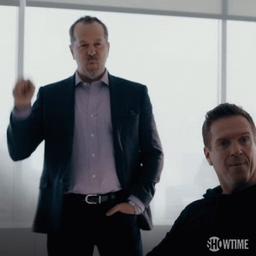 The image shows two men dressed in suits standing next to each other with one of them making a gesture or giving an indication. The man wearing the suit appears to be gesturing towards another man who is sitting down, possibly indicating that he wants to engage him in conversation or discuss something important. The scene takes place near a window, suggesting they might be in a professional setting like a conference room or office.