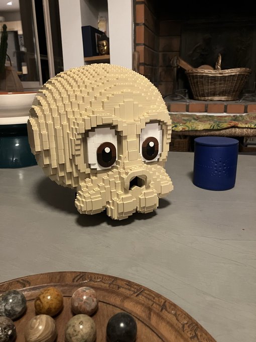 A large LEGO sculpture of a tan skull with big round eyes sits on a gray surface next to a blue candle holder, with a wooden tray of polished stones in the foreground.