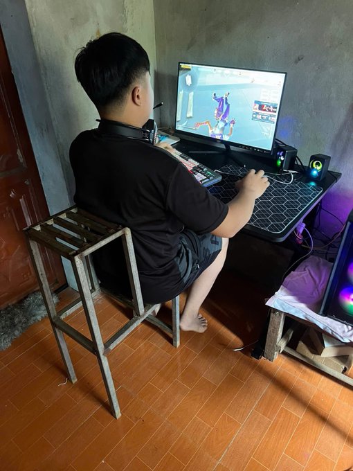 A young man wearing a black t-shirt sits barefoot on a simple wooden chair at a gaming desk, playing a video game on a monitor displaying a colorful character.