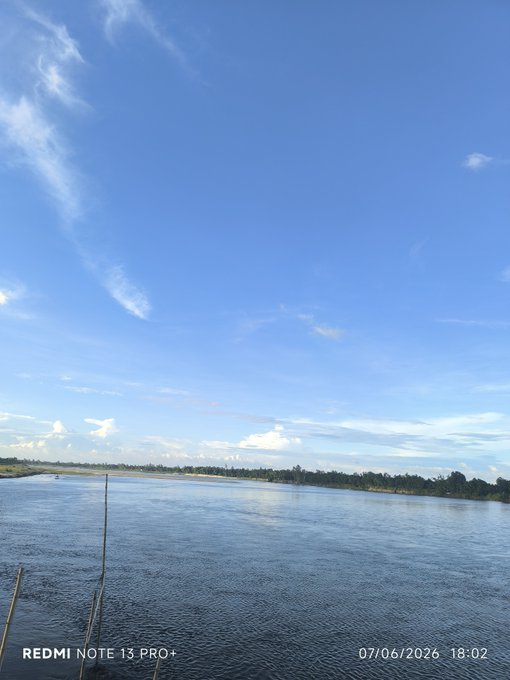 A scenic view of a calm river or lake with a distant shoreline under a blue sky with wispy clouds, timestamped 07/06/2026 18:02, taken on a REDMI NOTE 13 PRO+.