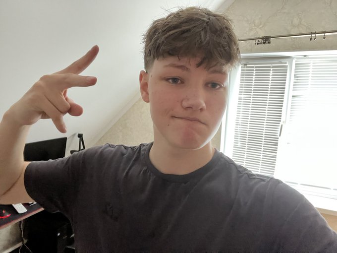 A young man in a dark gray t-shirt taking a selfie indoors near a window with blinds, pointing his finger upward.