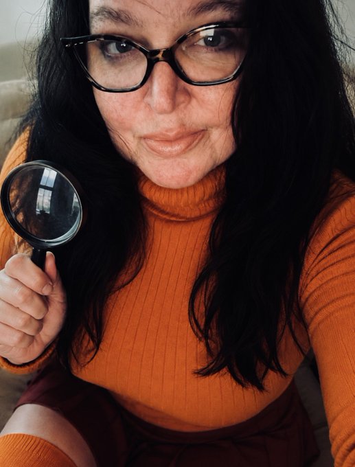A selfie-style photo of a woman with dark hair, glasses, and an orange ribbed turtleneck sweater holding a magnifying glass up to the camera.