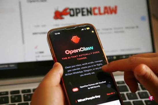 A person holds a smartphone displaying the 'OpenClaw' app website with the tagline 'THE AI THAT ACTUALLY DOES THINGS,' shown in front of a laptop with the same site open.