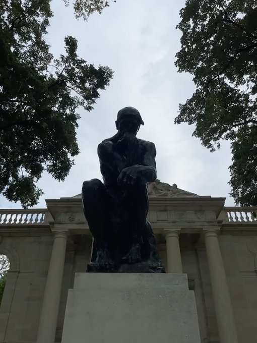 A bronze sculpture of The Thinker by Auguste Rodin, positioned on a pedestal in front of a classical building with columns, framed by tree branches against a cloudy sky.