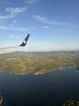 The image shows an aerial view of a body of water, possibly a lake or ocean, with a large airplane wing visible above it. The sky surrounding the water has some clouds, adding depth to the scene. The photo appears to be taken from inside an airplane window, capturing the beauty and vastness of the landscape below.