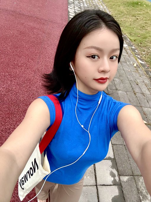 A young woman takes a selfie on a red running track, wearing a bright blue sleeveless turtleneck top, white earphones, and a tote bag with the text 'NOW IS NOT' visible.