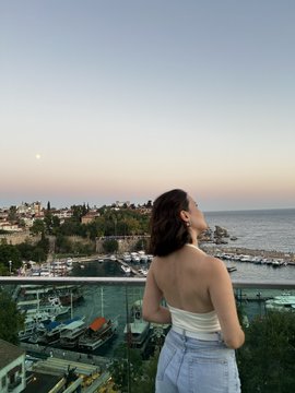 A photograph of a woman with dark hair tied back, wearing a white tube top and blue jeans, leaning on a balcony railing and looking out toward a marina and sunset/dusk sky.