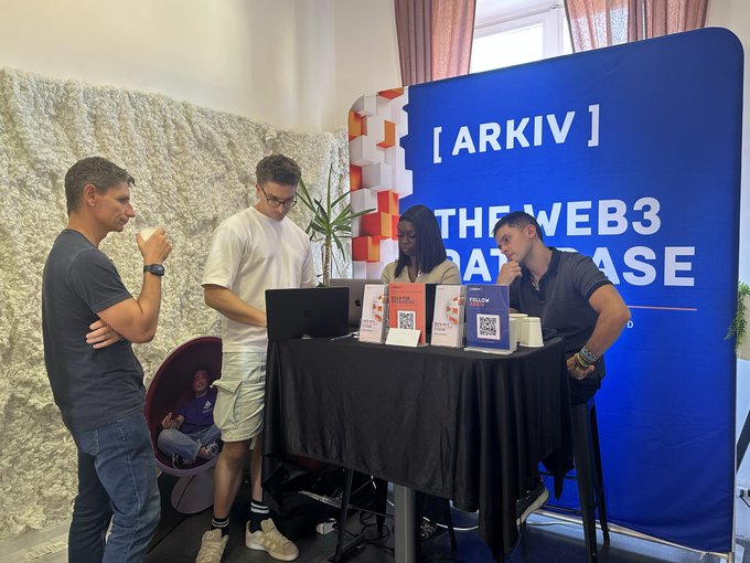 Four people gathered around a table at a booth with a blue '[ARKIV] THE WEB3 DATABASE' banner, with QR codes and 'FOLLOW' signs displayed on the table.