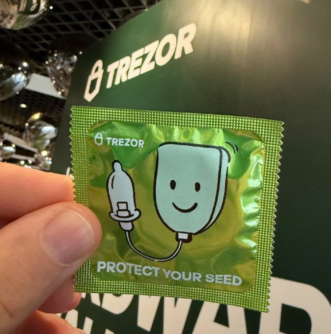 A hand holds a green Trezor-branded condom wrapper with the text 'PROTECT YOUR SEED' and a cartoon character, set against a Trezor-branded backdrop.