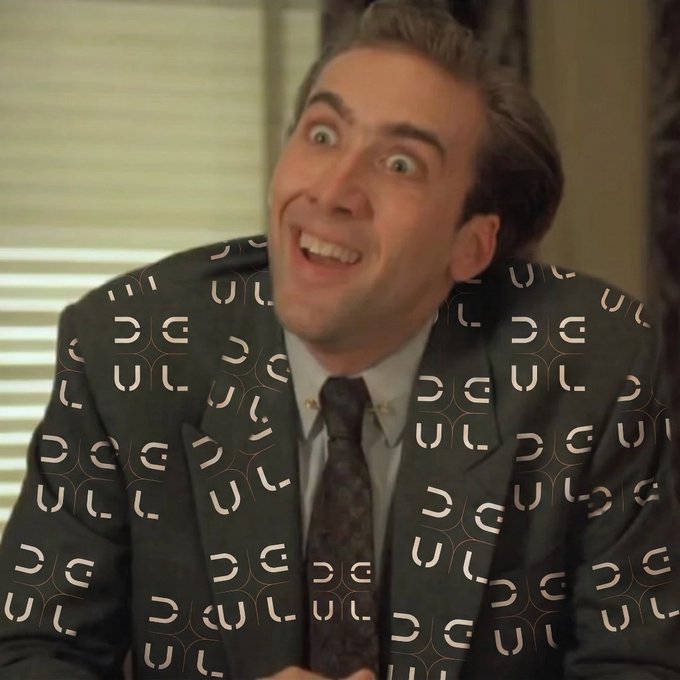 The image features a man wearing a suit with a patterned jacket and tie. The man has his mouth open, making an odd or funny face as he looks at something off-camera. The background of the image shows a window, suggesting that they might be in an office setting.