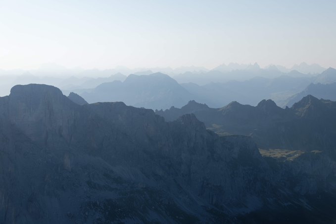 A scenic landscape photograph showing layers of mountain ridges fading into the distance under a pale blue sky, with sharp rocky peaks in the foreground and a hazy atmospheric perspective.
