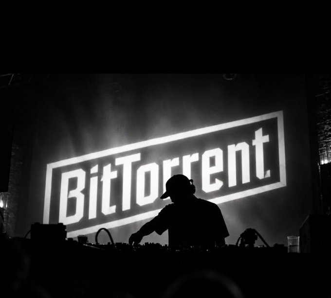 A black-and-white photograph shows the silhouette of a DJ wearing headphones and a cap, working at equipment with a large 'BitTorrent' logo illuminated on a screen behind them.