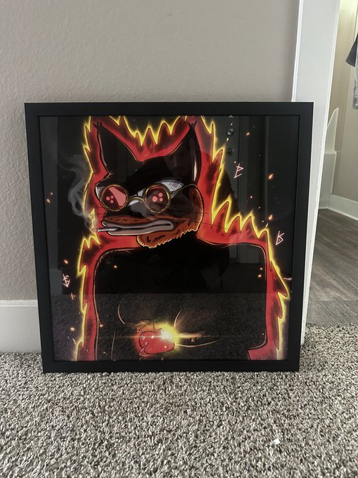 A framed artwork of a stylized cat wearing goggles and a hat, surrounded by fiery flames, leans against a wall on a carpeted floor with its reflection visible in the glass.