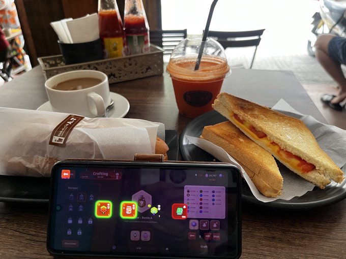 A smartphone displaying a crafting menu game screen rests on a wooden table next to a plate with a grilled cheese sandwich, an iced drink with a straw, a coffee cup, and a wrapped food item.