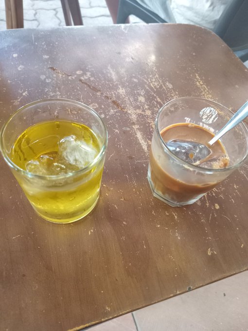 Two glasses sit on a worn wooden table — a yellow iced drink on the left and a brown coffee or iced latte with a spoon on the right.