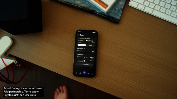 A smartphone on a wooden desk displays a GalaxyOne app screen showing a Cash account balance of $10,008.76, illustrating the post's mention of depositing $10,000 cash into the platform. The image's disclaimer notes "Actual GalaxyOne account shown. Paid partnership. Terms apply," aligning with the user's reference to a paid promotional opportunity.