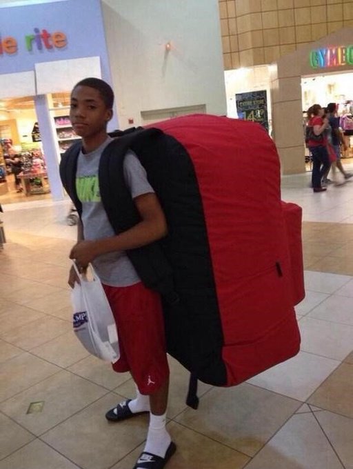 A young man in a mall carries an oversized red backpack so large it nearly swallows him, with stores like "rite" and "Game" visible in the background. The humor plays on the post's boast about Backpack "revolutionizing finance," mocking the project by exaggerating the literal backpack to comically oversized proportions.