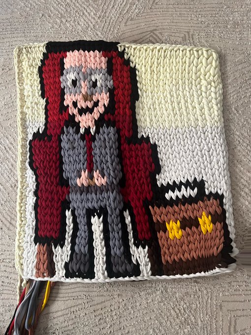 A handmade knitted or crocheted textile art piece depicting a pixelated figure with a beard in a red cloak standing next to a small brown creature with yellow eyes.