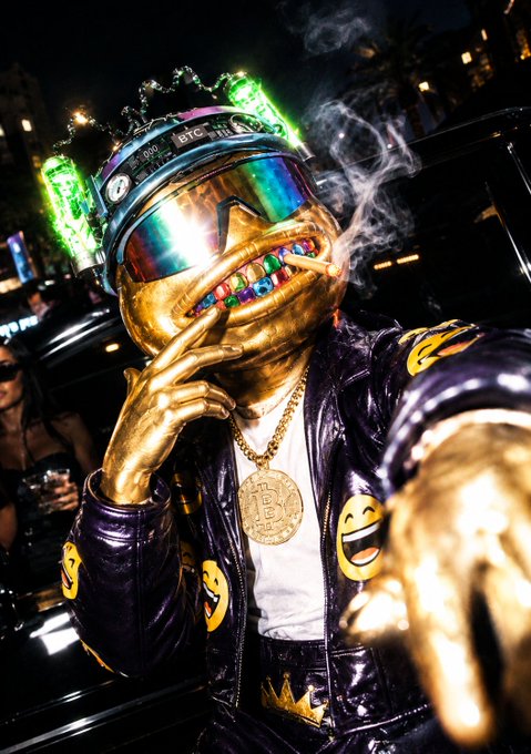 A gold-painted person wearing a reflective helmet, jeweled mouth guard, Bitcoin necklace, and purple smiley-face jacket poses at night.