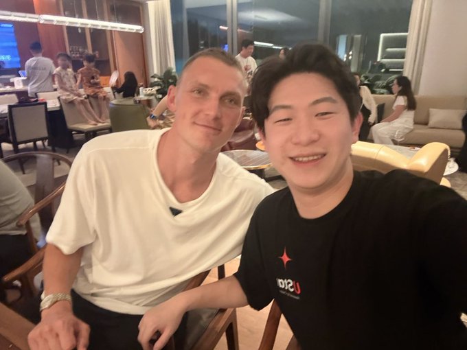 Two men take a selfie together in what appears to be an upscale lounge or hotel lobby, with one wearing a black t-shirt featuring red text/logo and the other in a white t-shirt, while other guests can be seen seated in the background. The image relates to the post as it shows the moment the user met Viktor Axelsen, the Danish badminton superstar, who is now learning about Bitcoin.