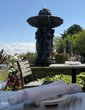 A stone fountain featuring gargoyle-like figures sits in a sunlit garden beside a dining table set with napkins and glassware. The image matches the post, as the table is positioned directly in front of the carved statues, overlooking the scenic view.