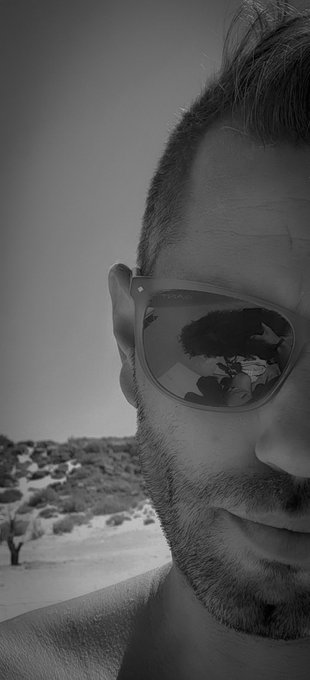 A black-and-white close-up selfie of a bearded man wearing sunglasses, which reflect a person standing near trees in an arid, desert-like landscape.