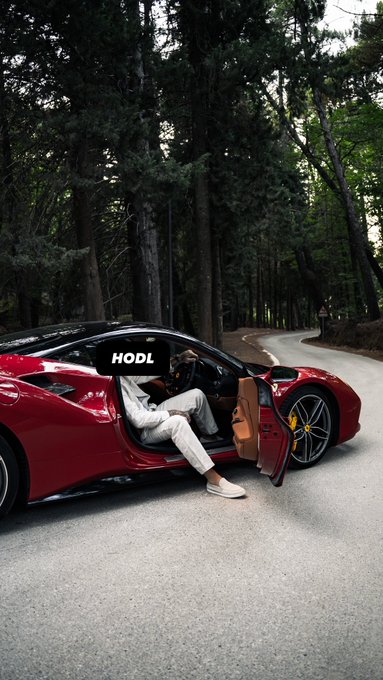 A man in a white outfit sits in the open door of a red sports car parked on a forest road, holding up a sign that reads 'HODL'.