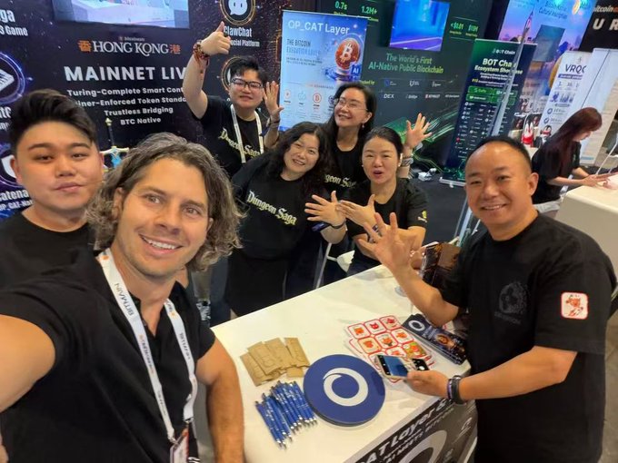 A group selfie taken at a Hong Kong conference booth, showing several smiling people wearing 'Dragon Saga' and 'Bitcoin' themed shirts, promoting 'MAINNET LIVE' with Bitcoin and BRC20 themes.
