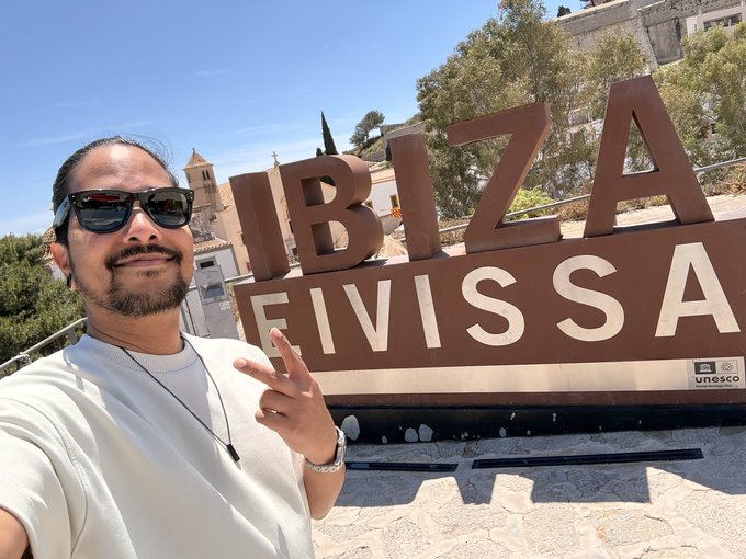 A man takes a selfie in front of the large "IBIZA EIVISSA" sign, marked with a UNESCO World Heritage plaque, flashing a peace sign on a sunny day. The "Gm" greeting fits perfectly with the cheerful, travel-vibe vibes of this iconic Balearic island landmark.