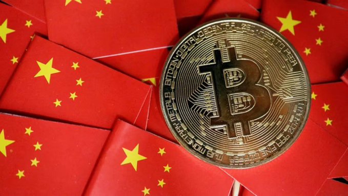 A gold Bitcoin token, inscribed with "BITCOIN DIGITAL DECENTRALIZED PEER TO PEER" and "COPPER FINE COPPER MJB MONETIZED," rests atop a scattered pile of small Chinese flags, symbolizing the reported plan to unban cryptocurrency in China by the end of 2026.