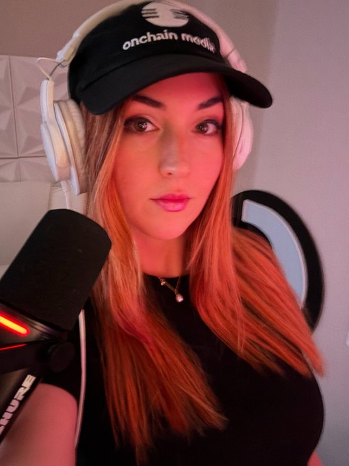 A young woman wearing an 'onchain media' cap and white headphones sits near a microphone with red lighting in the background.